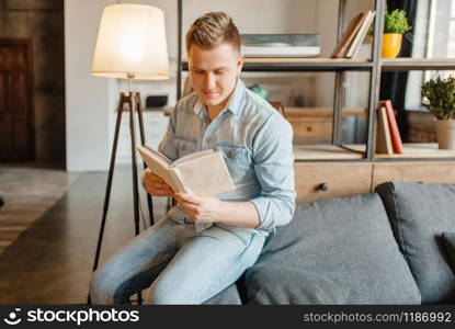 Young man sitting in couch and reading a book. Male person reads in living room, floor lamp and shelf on background. Young man sitting in couch and reading a book