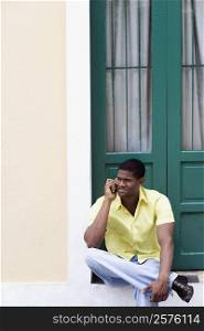 Young man sitting in front of a door and talking on a mobile phone