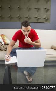 Young man sitting in front of a laptop and writing on a spiral notebook