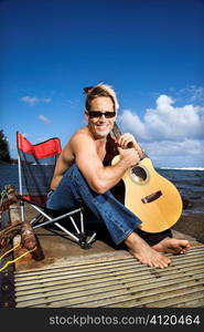 Young Man Sitting Lakeside and Holding Guitar