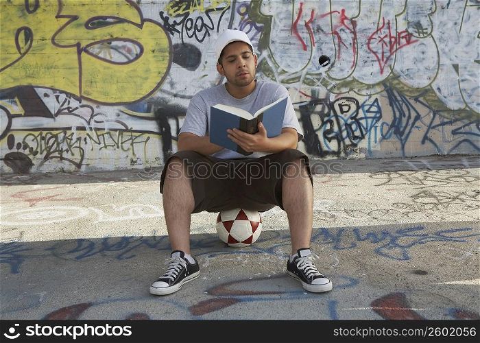 Young man sitting on a soccer ball and reading a book
