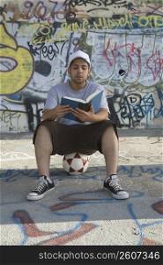 Young man sitting on a soccer ball and reading a book