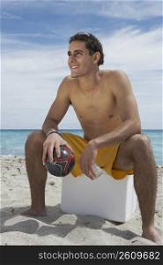 Young man sitting on an ice box on the beach and holding a soccer ball