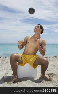 Young man sitting on an ice box on the beach and playing with a soccer ball