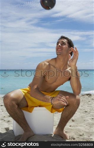 Young man sitting on an ice box on the beach and playing with a soccer ball