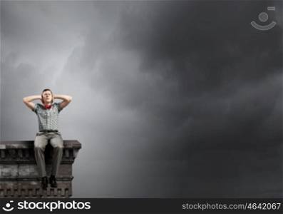 Young man sitting on building top and covering ears with hands
