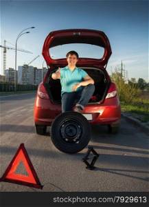 Young man sitting on car trunk and holding foot on spare wheel