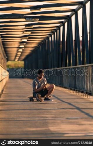 Young man sitting on his skateboard outside on a pedestrian bridge by the river, texting.