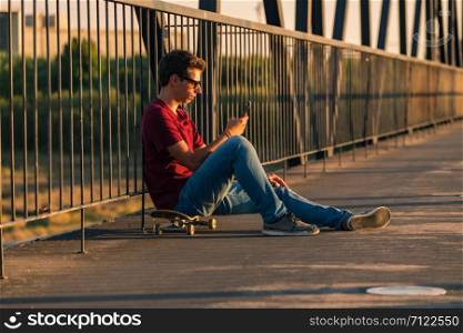 Young man sitting on his skateboard outside on a pedestrian bridge by the river, texting.