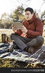 Young man sitting on picnic blanket in forest looking at digital tablet