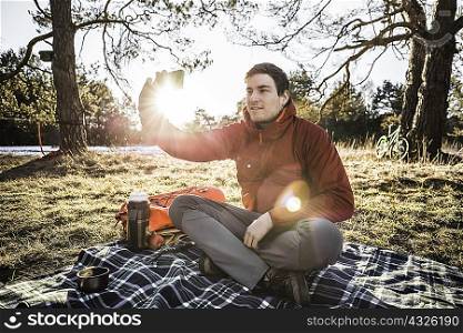 Young man sitting on picnic blanket in forest taking smartphone selfie