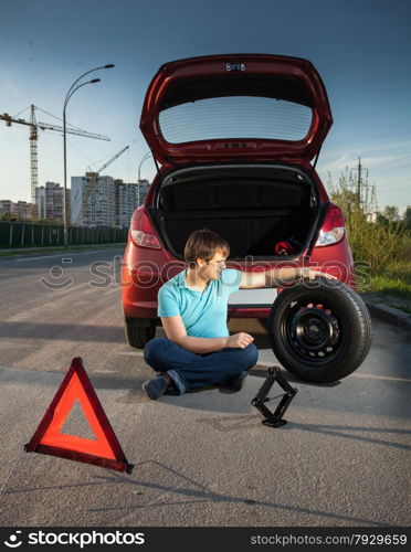 Young man sitting on road and leaning against broken car