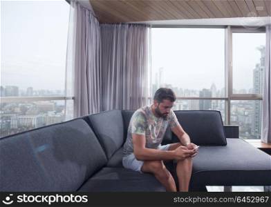 young man sitting on sofa and using a mobile phone near the window at home