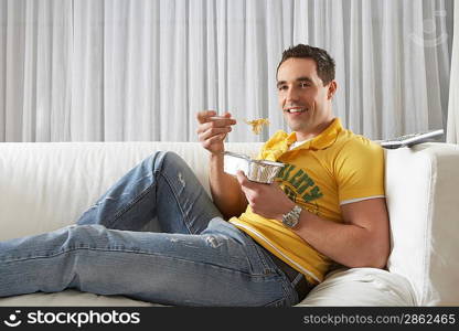 Young man sitting on sofa eating noodles from takeaway tray