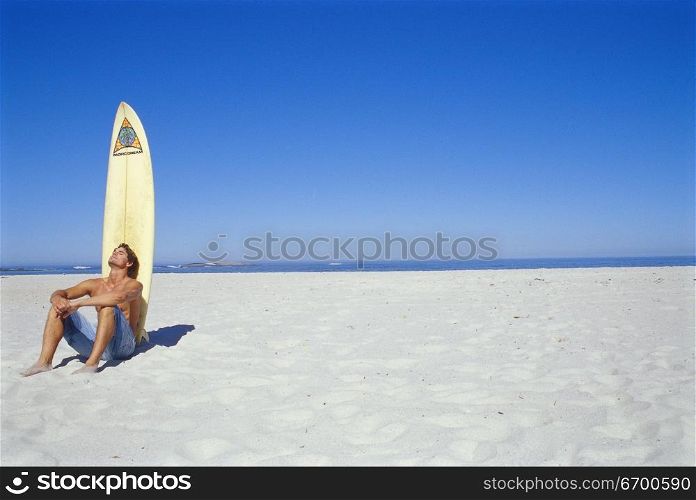 Young man sitting on the beach in front of a surfboard