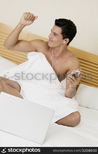 Young man sitting on the bed and listening to an MP3 player with a laptop in front of him