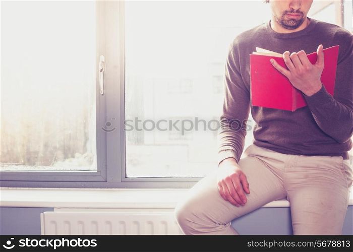 Young man sitting on window sill and reading a red book