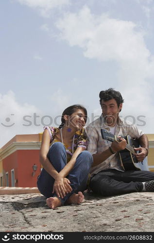 Young man sitting with a young woman and playing a guitar
