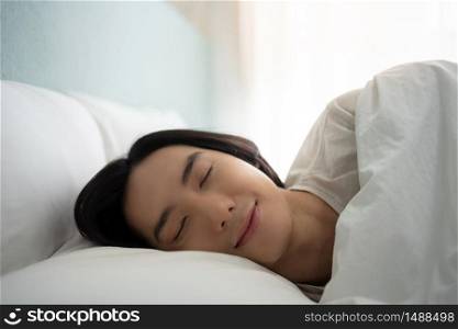Young man sleeping well and dreaming in white bed in the morning on holiday. Happy smiling Asian male enjoying lying on comfortable soft bedding, pillow, blanket and mattress in bedroom at home. eyes closed.