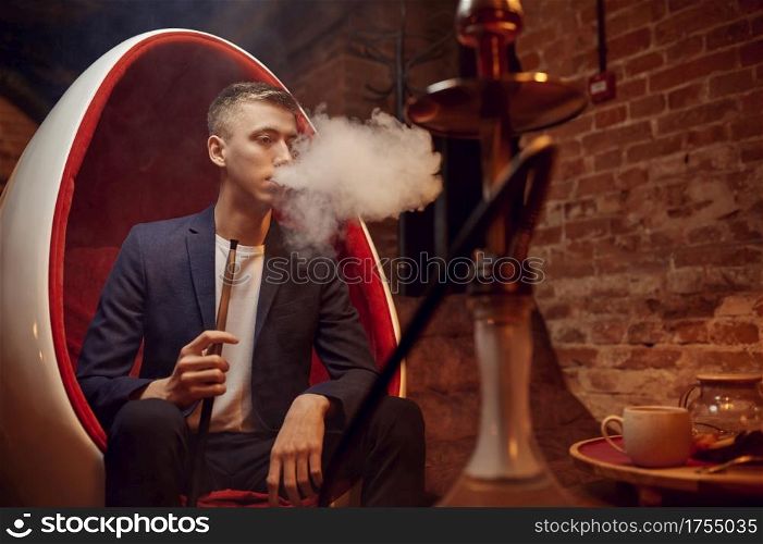 Young man smokes in hookah bar, hooka chill out. Shisha smoking, traditional bong culture, tobacco aroma for relaxation, rest with hooka. Young man smokes in hookah bar, hooka chill out