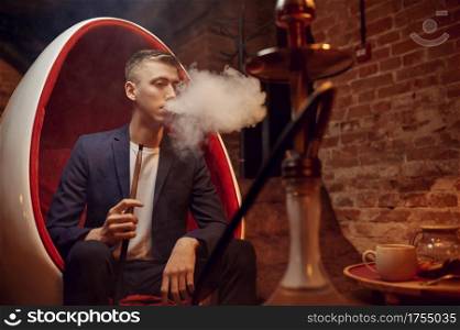 Young man smokes in hookah bar, hooka chill out. Shisha smoking, traditional bong culture, tobacco aroma for relaxation, rest with hooka. Young man smokes in hookah bar, hooka chill out