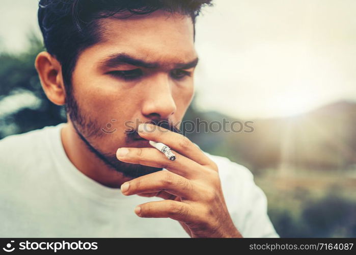 Young man smoking cigarette in the outdoors and nature background. Selective focus at cigarette.