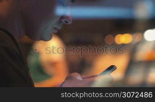Young man spending evening with smart phone in cafe. He communicating in social network service or typing sms. View through the glass