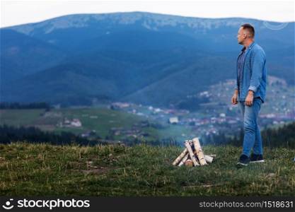 Young man standing by a burning fire under the evening sky in the mountains. Active lifestyle concept.. Young man standing by a burning fire under the evening sky in the mountains. Active lifestyle concept