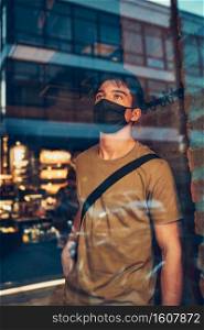 Young man standing in coffee shop at store front in the city center in the evening, wearing the face mask to avoid virus infection and to prevent the spread of disease in time of coronavirus