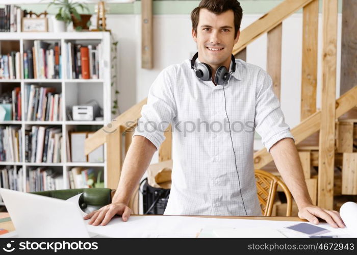 Young man standing in creative office. Smiling young designer standing in creative office in front of his desk