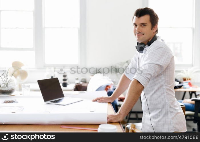 Young man standing in creative office. Smiling young designer standing in creative office in front of his desk