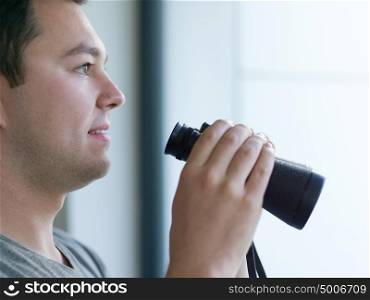 Young man standing looking through a glass window with binoculars as he watches something in the distance