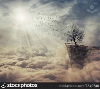 Young man standing on the peak of a cliff over clouds watching at a flock of birds flying from a strange, bare tree. The tree of death symbol, journey and discover.