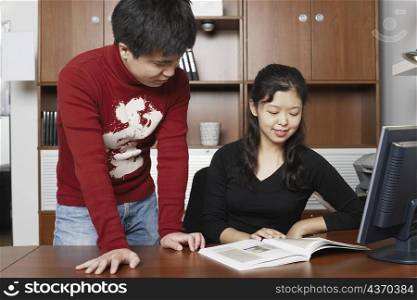 Young man standing with a mid adult woman sitting beside him reading a book