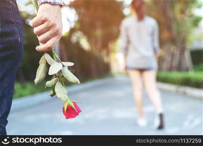 young man standing with a Red Rose on Hand Sadness Love in Ending Break up of Relationship Blurred woman Back Side Walking away parting in public park outdoor. Broken Heart in Valentine day Concept.