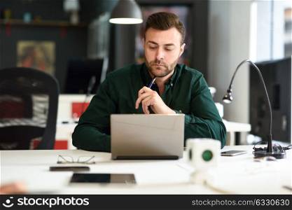 Young man studying with laptop computer on white desk. Attractive guy with beard wearing casual clothes.