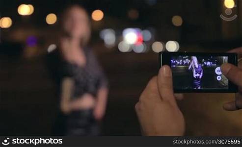 Young man takes photos of his girlfriend on the city streets at night. Close up of male hands holding cellphone. Shallow depth of field. Focus on image in phone then focus on girl.