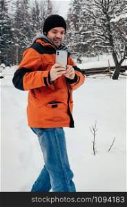 Young man taking picture with his smartphone, smiling. Blue jeans, orange garment .Winter day, snow hill, trees, countryside. Fresh air. Fun and leisure concept