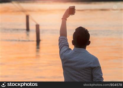 young man taking selfie picture using his smartpone, enjoying sunset time outdoors.
