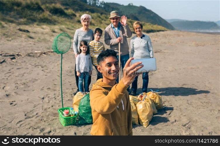 Young man taking selfie with mobile to group of volunteers after cleaning the beach. Selective focus on guy in foreground. Young man taking selfie to group of volunteers after cleaning beach