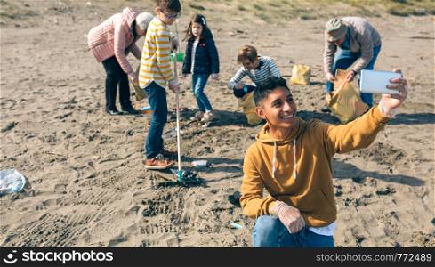 Young man taking selfie with mobile to group of volunteers cleaning the beach. Selective focus on guy in foreground. Young man taking selfie to group of volunteers cleaning beach