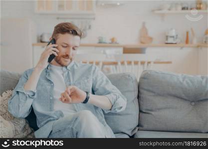 Young man talking on phone while checking time on his wristwatch, sitting on couch in his apartment with crossed legs, agreeing on meeting with friend, choosing best time to meet. Young man talking on phone with his friend while sitting on couch