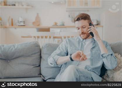 Young man talking on phone while checking time on his wristwatch, sitting on couch in his apartment with crossed legs, agreeing on meeting with friend, choosing best time to meet. Young man talking on phone with his friend while sitting on couch