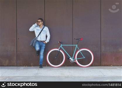 Young man talking on smart phone while standing by bicycle against metallic wall at sidewalk in city