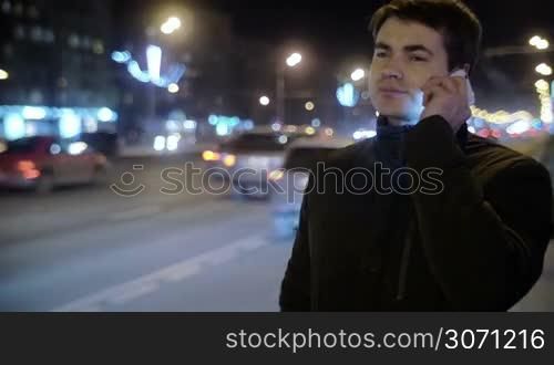 Young man talking on the phone outdoor at night. He standing near the busy road, cars passing by, city lights in background