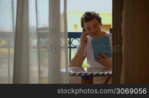 Young man talking on the phone with tablet PC in hands on hotel balcony