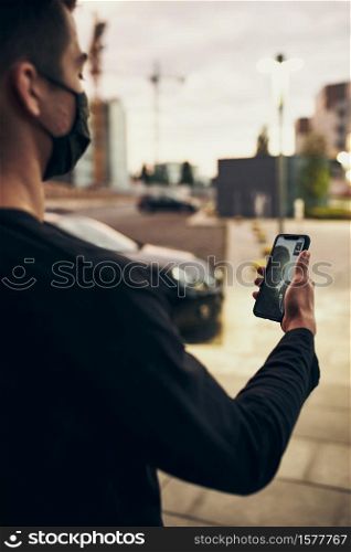 Young man talking on video call standing outdoors in the street in the evening wearing the face mask to avoid virus infection and to prevent the spread of disease in time of coronavirus