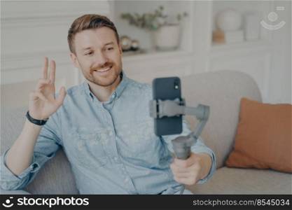Young man talking with his friend through online video chat using telephone attached to gimbal, waving hello with hand while smiling, happy after not seeing each other for long time. Young man talking with his friend through online video chat using telephone