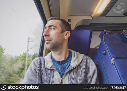Young man traveling on a train and looks out the window. A blue suitcase is next to him.
