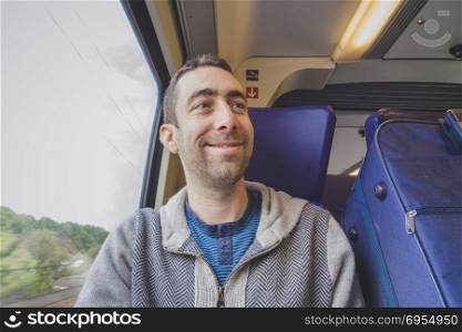 Young man traveling on a train and smiling at someone. A blue suitcase is next to him.